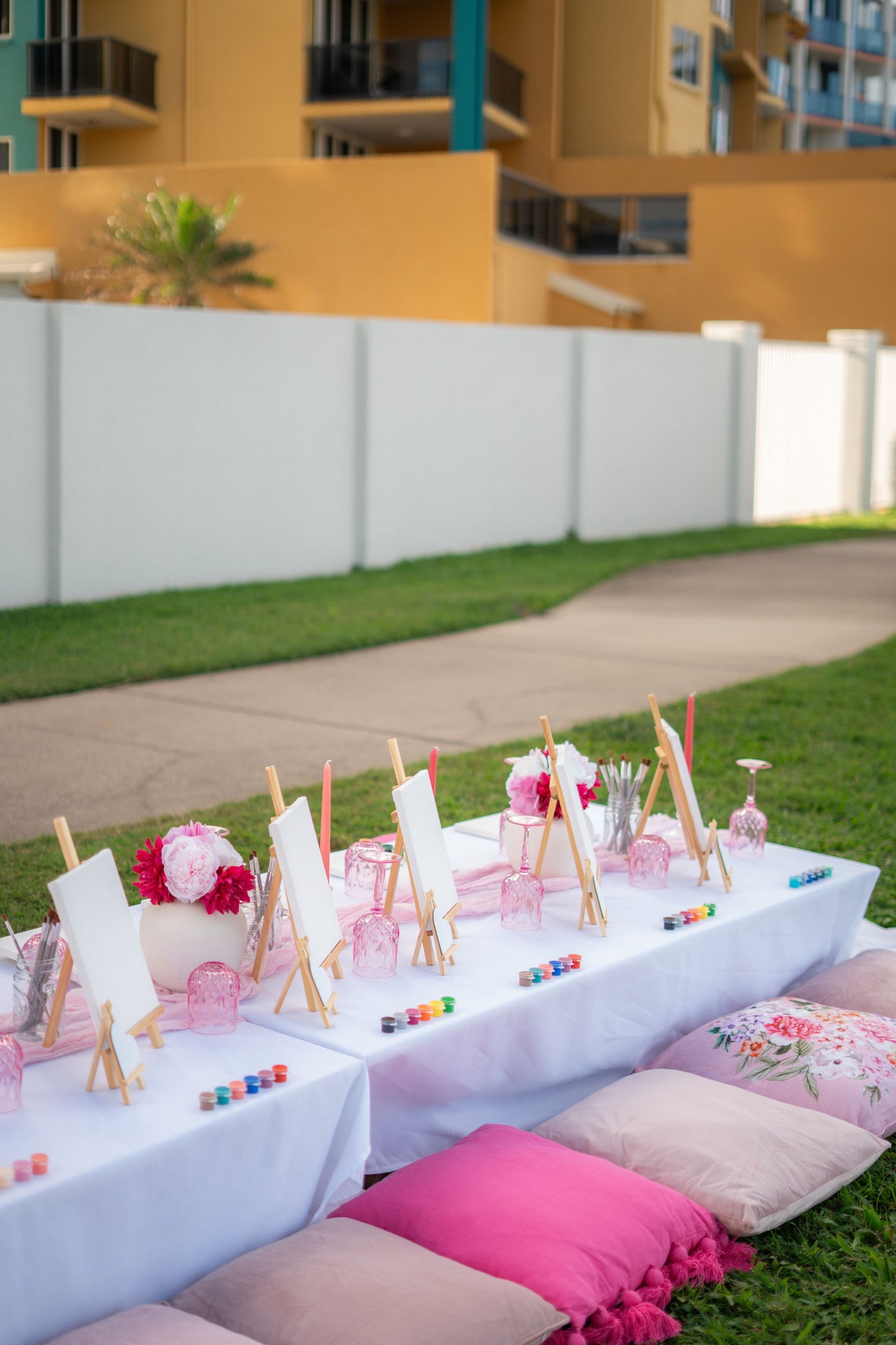Outdoor setting with a table decorated for a birthday party, featuring pink and white chairs, colourful decorations, and pink pillows on the grass.