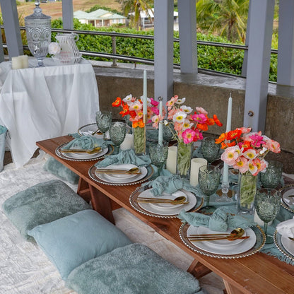 A set luxury picnic table with plates, silverware, colorful floral arrangements, and decorative pillows on a balcony with a tropical landscape in the background.