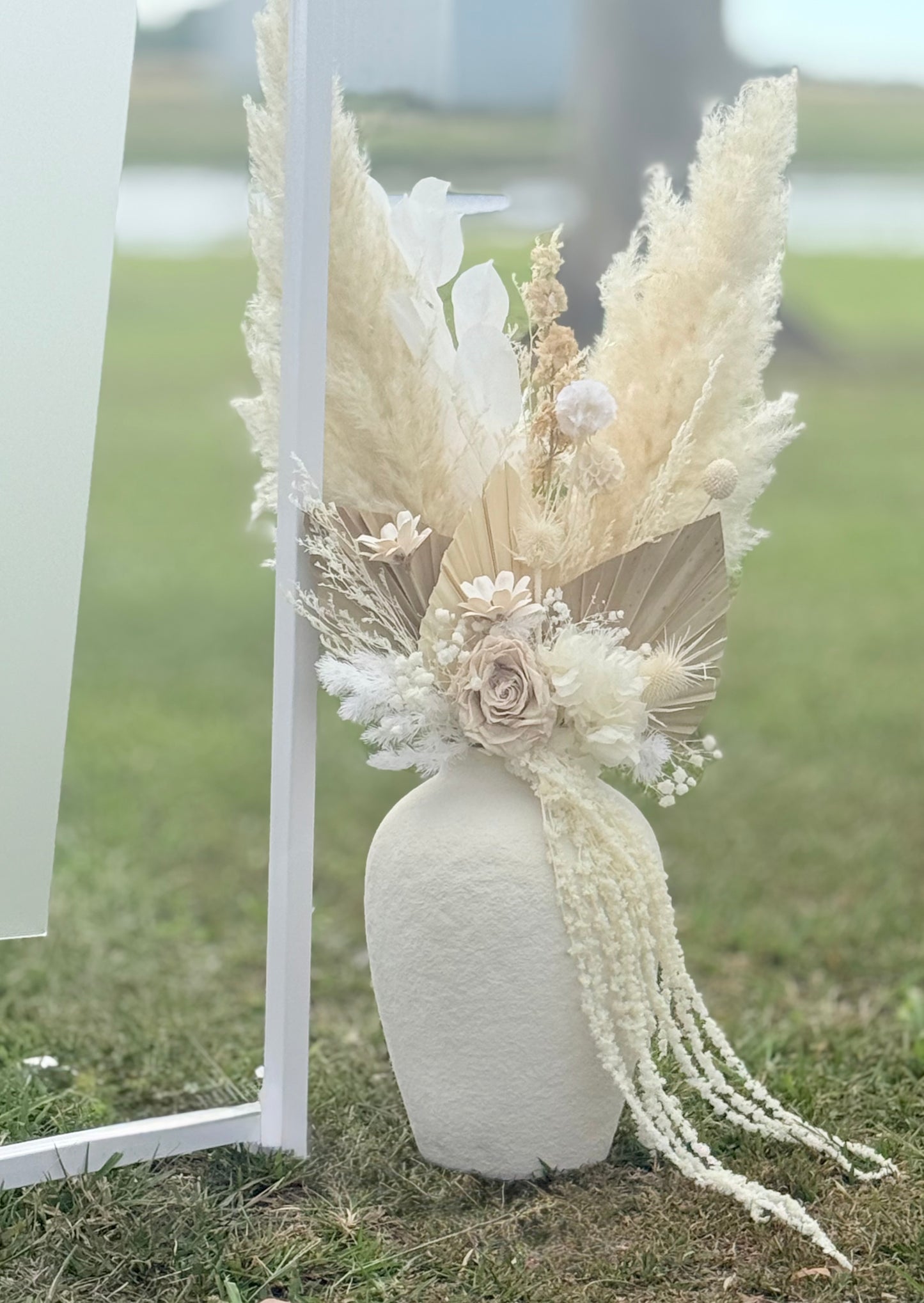 Decorative floral arrangement with pampas grass and roses in a white vase on grass.