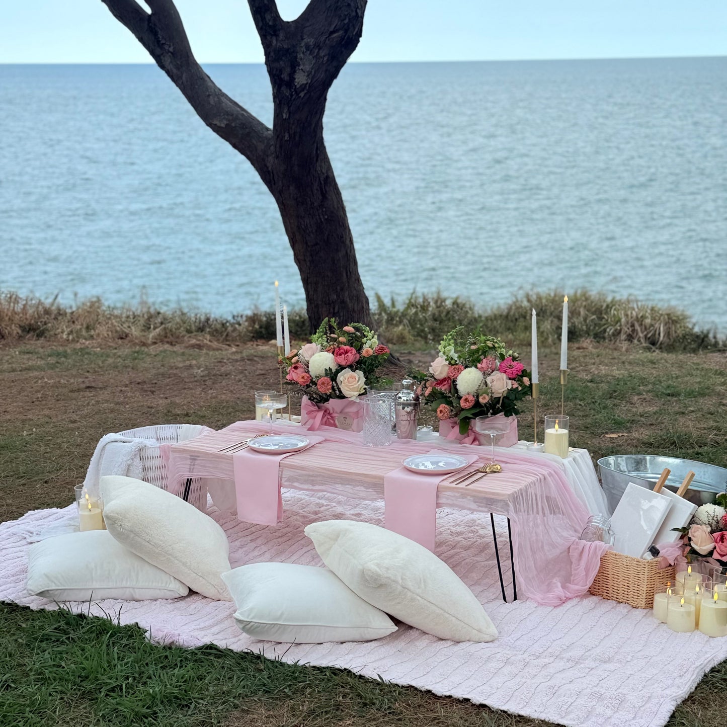 Picnic setup with pink tablecloth, flowers, and candles by a body of water.