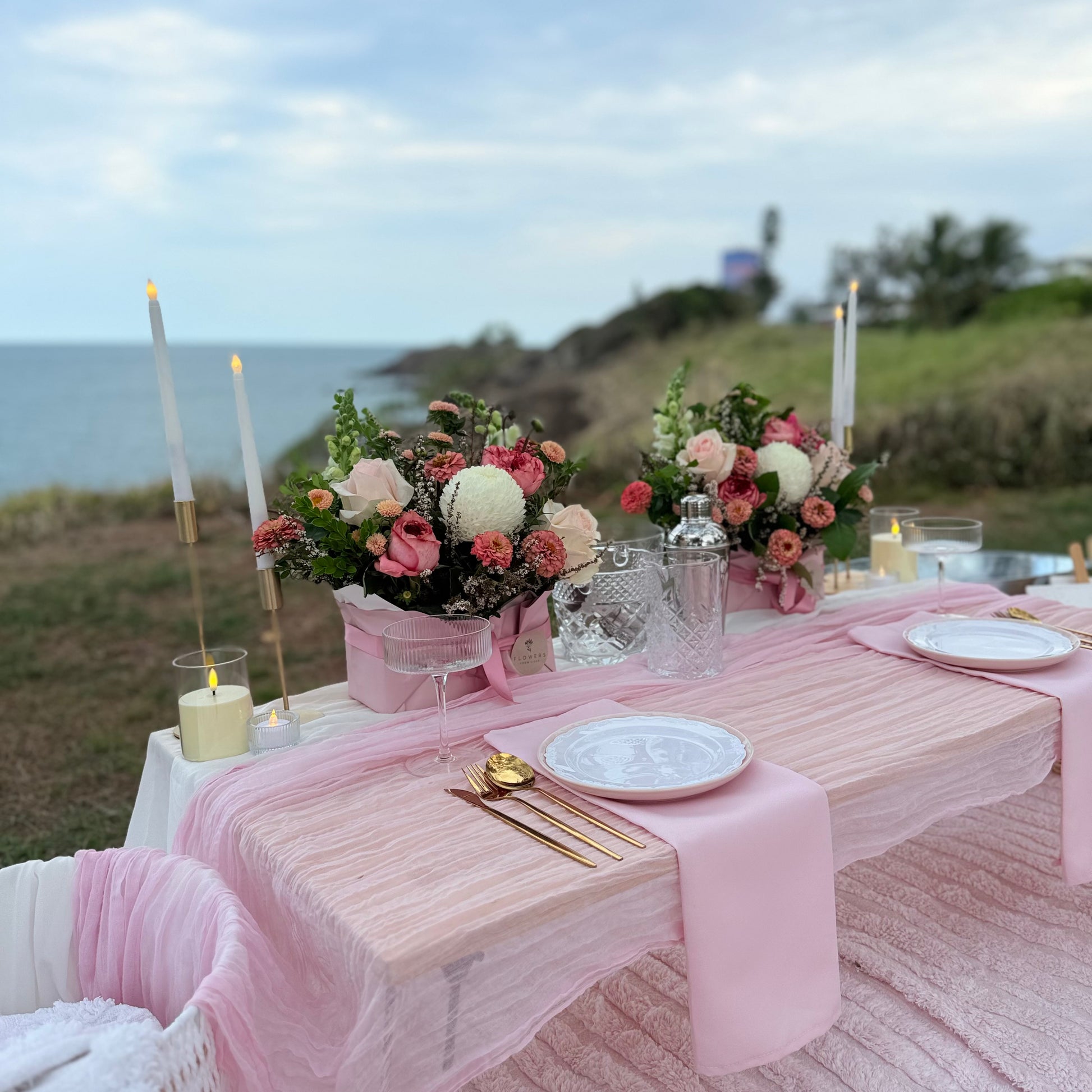 Decorated outdoor table with pink tablecloth, flowers, and candles by the ocean.