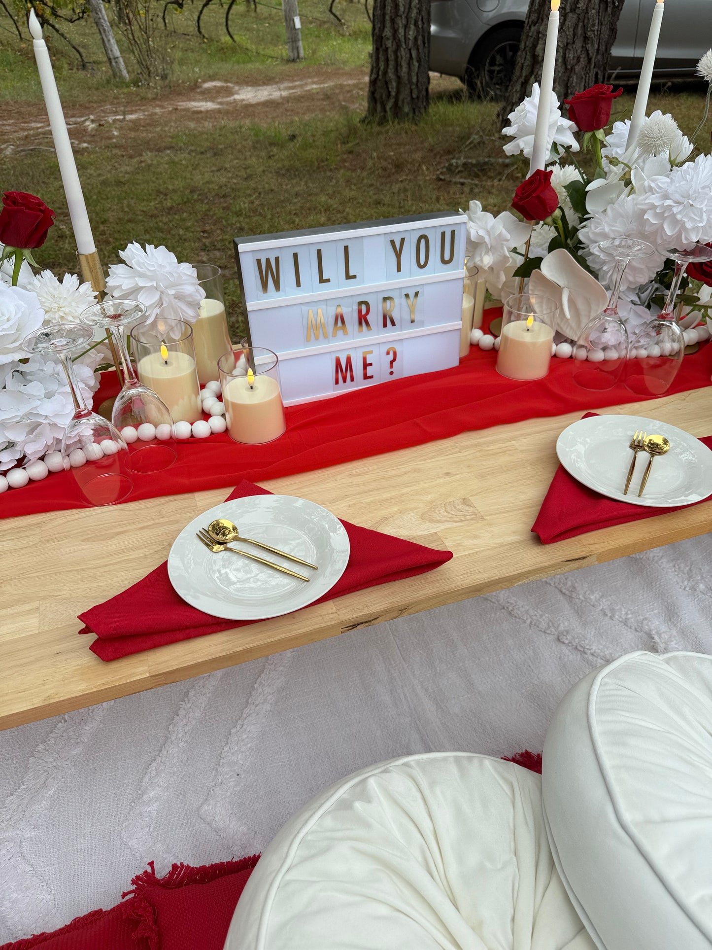 Decorated table with 'WILL YOU MARRY ME?' sign, candles, and flowers on a red tablecloth.