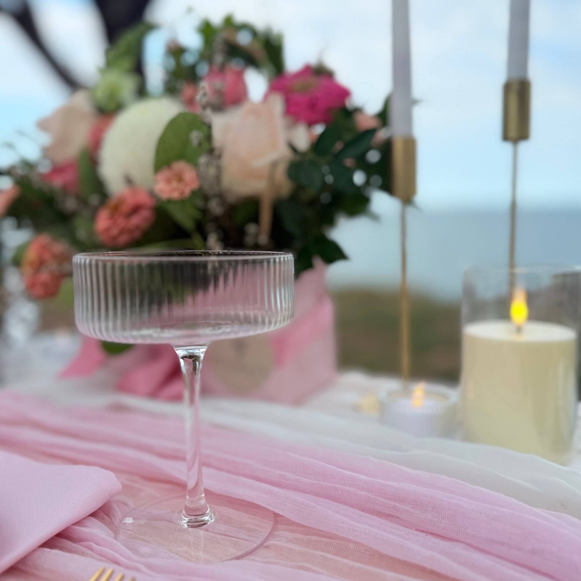 Clear glass coupe on a table with pink tablecloth and floral arrangement in the background