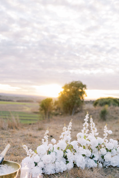 White floral arrangements in a dry grassy field at sunset, with trees and an open landscape in the background under a cloudy sky.