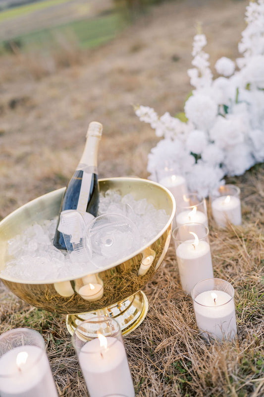 A gold champagne bowl filled with ice and a bottle of champagne, styled outdoors on dry grass with lit white candles and white flowers in the background.