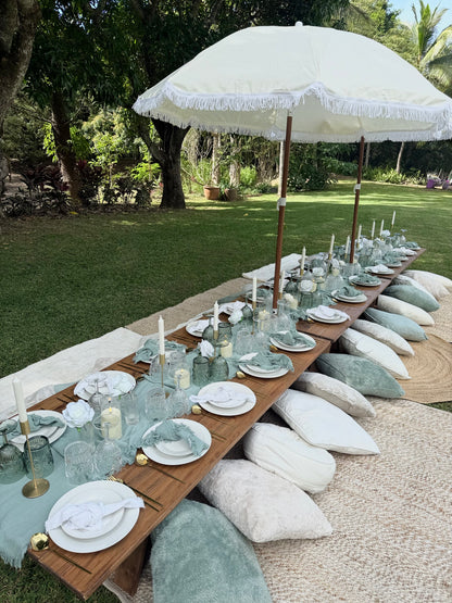Outdoor dining setup with a long wooden table, white plates, glasses, and a large white umbrella in a garden setting.