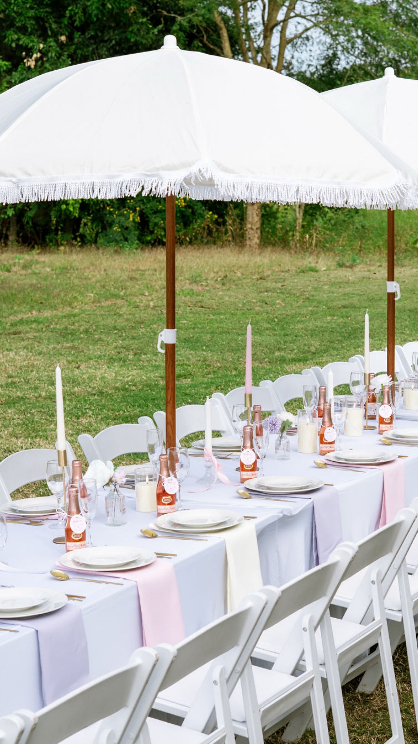Outdoor dining setup with white tablecloths, white chairs, and white umbrellas in a grassy area.