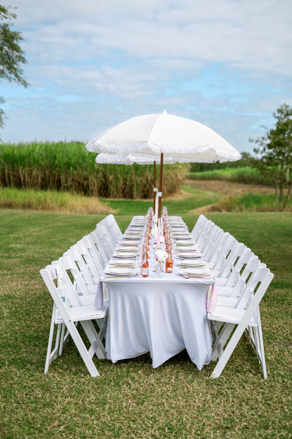 Dining table set with white tablecloth, chairs, and a large white umbrella outdoors on a grassy area.