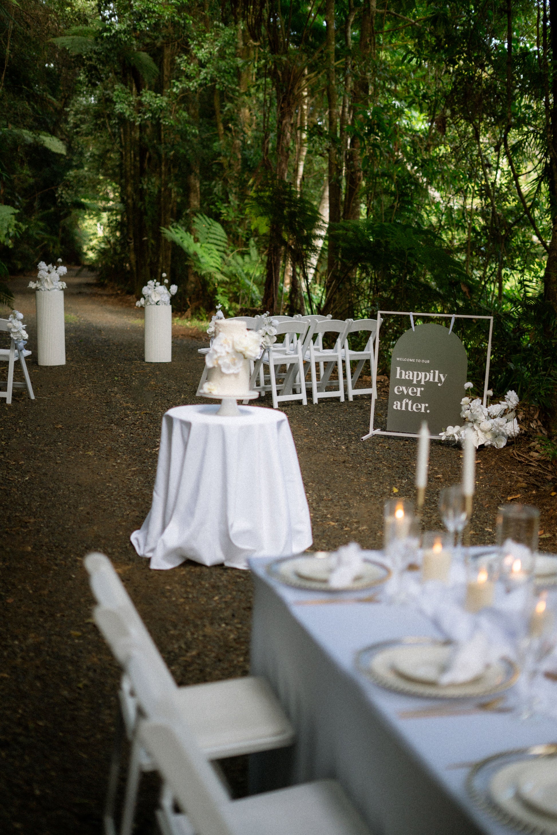 Decorated outdoor wedding setup with tables, chairs, and a 'Happily Ever After' sign in a forest.