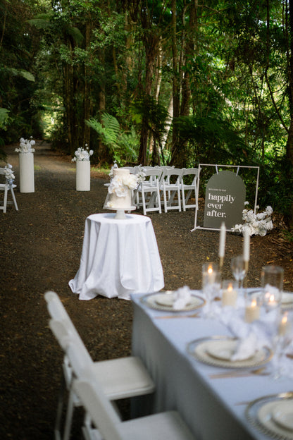 Decorated outdoor wedding setup with tables, chairs, and a 'Happily Ever After' sign in a forest.