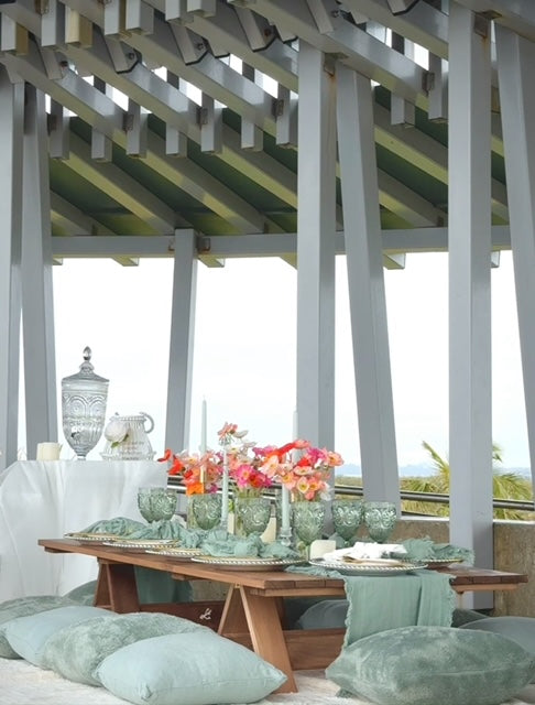 An elegantly set picnic table under a wooden pergola, featuring plush cushions, low trestle tables, and a drink station with a crystal decanter.