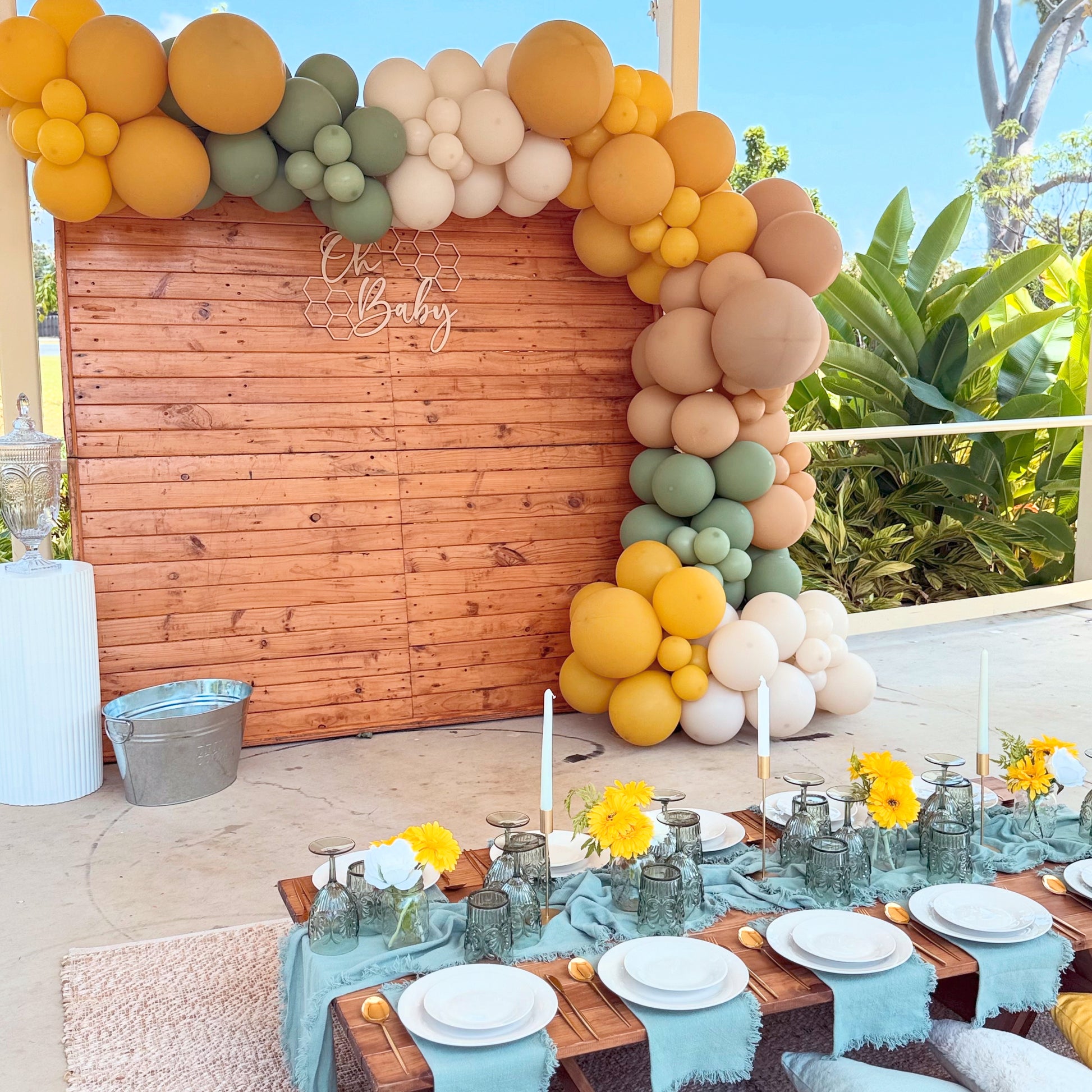 Decorative balloon arch with a wooden backdrop and a table set for a baby shower outdoors.