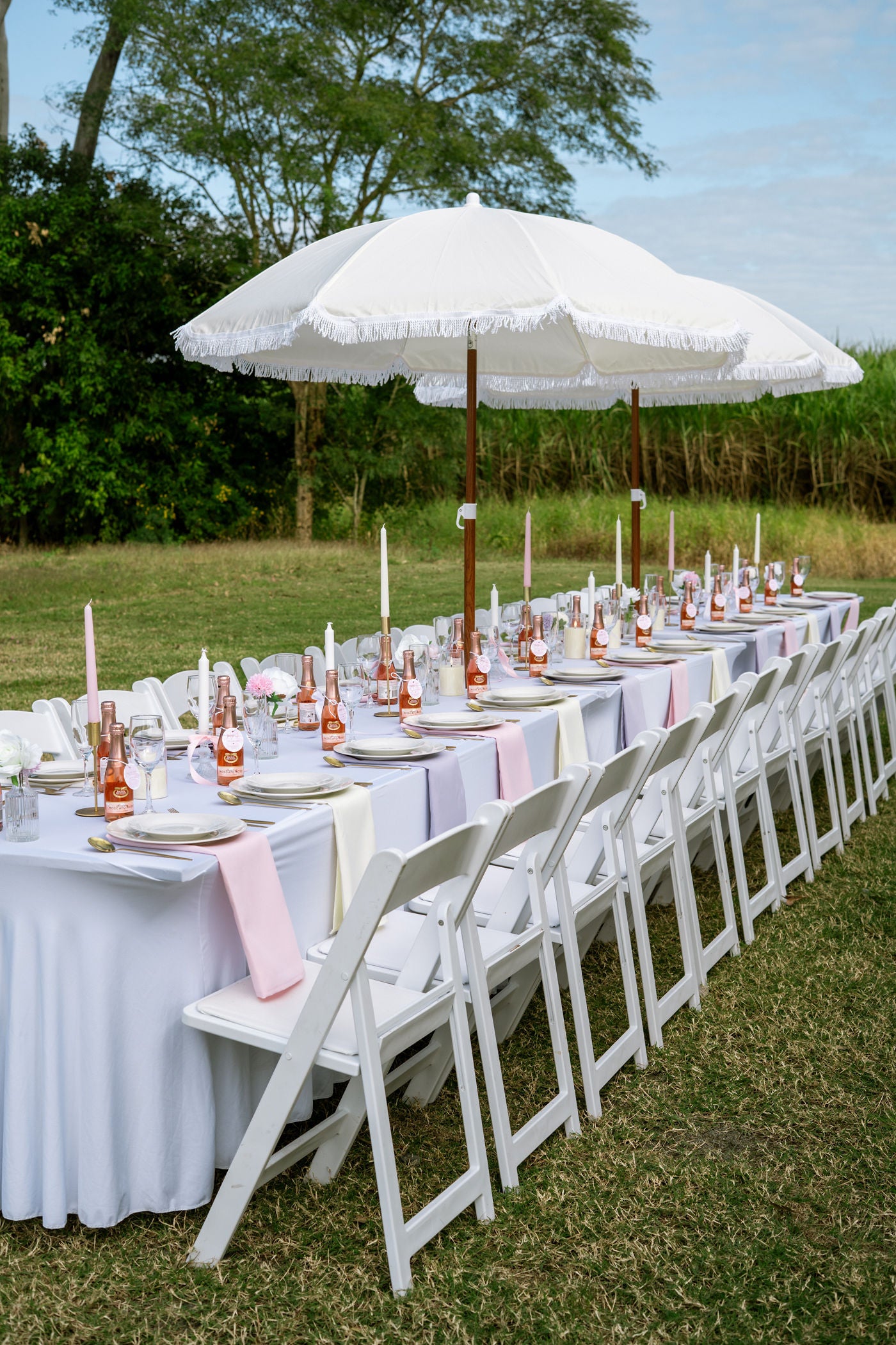 Outdoor wedding setup with white tables, chairs, and a large umbrella on a grassy field.