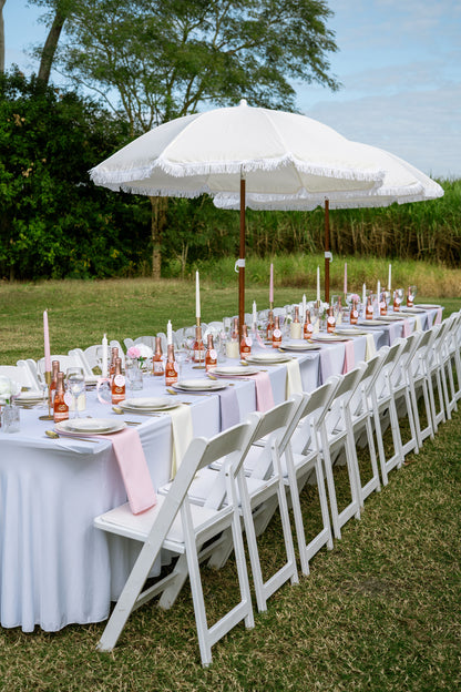 Outdoor wedding setup with white tables, chairs, and a large umbrella on a grassy field.