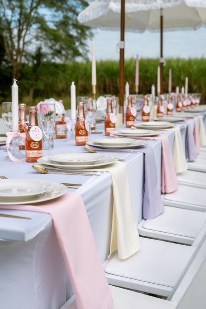 Outdoor table setting with pastel tablecloths, plates, and bottles under white umbrellas.