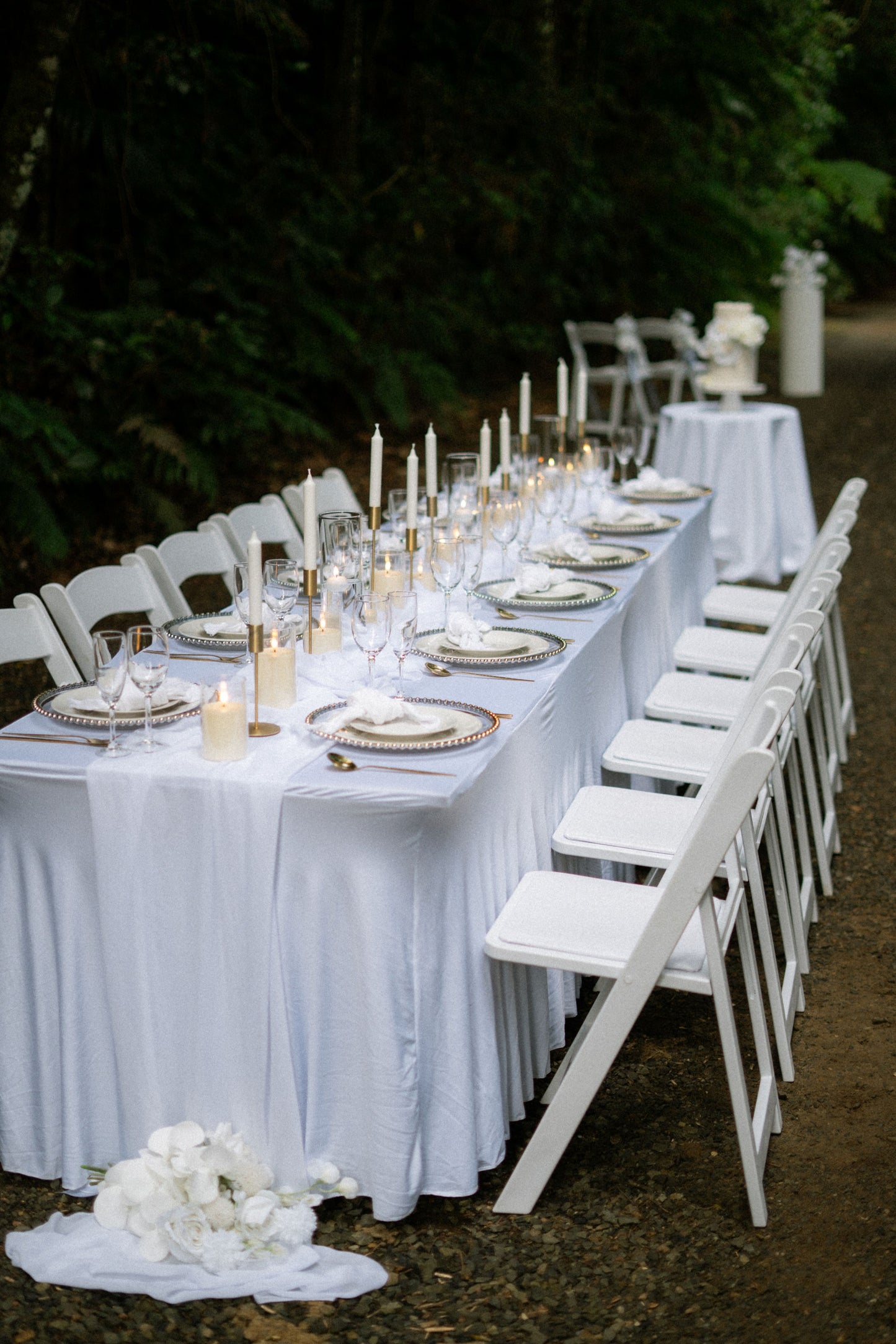 Long table set for a formal event with white tablecloth, plates, candles, and chairs outdoors.