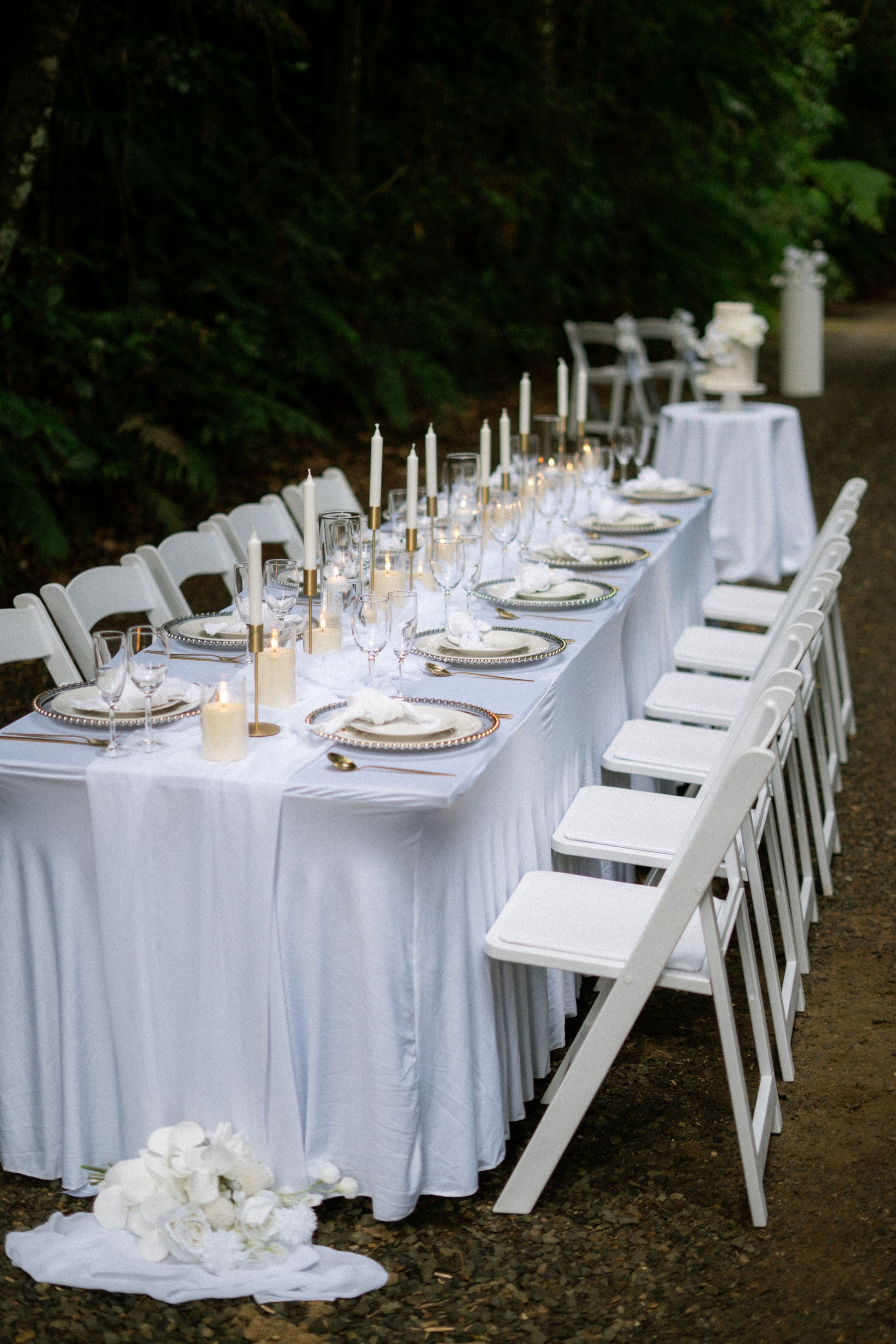 Long table set for a formal event with white tablecloth, plates, candles, and chairs outdoors.
