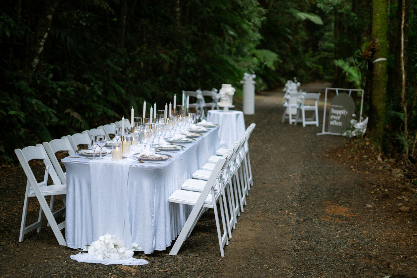 Long table set for a meal with white tablecloth and chairs in a forest setting.
