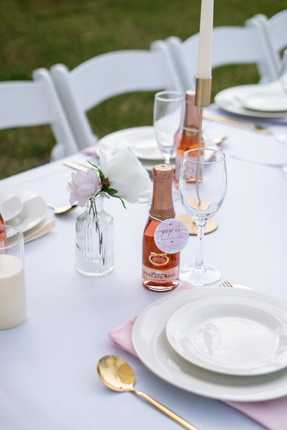 Table setting with bottles, glasses, and plates on a white tablecloth outdoors.