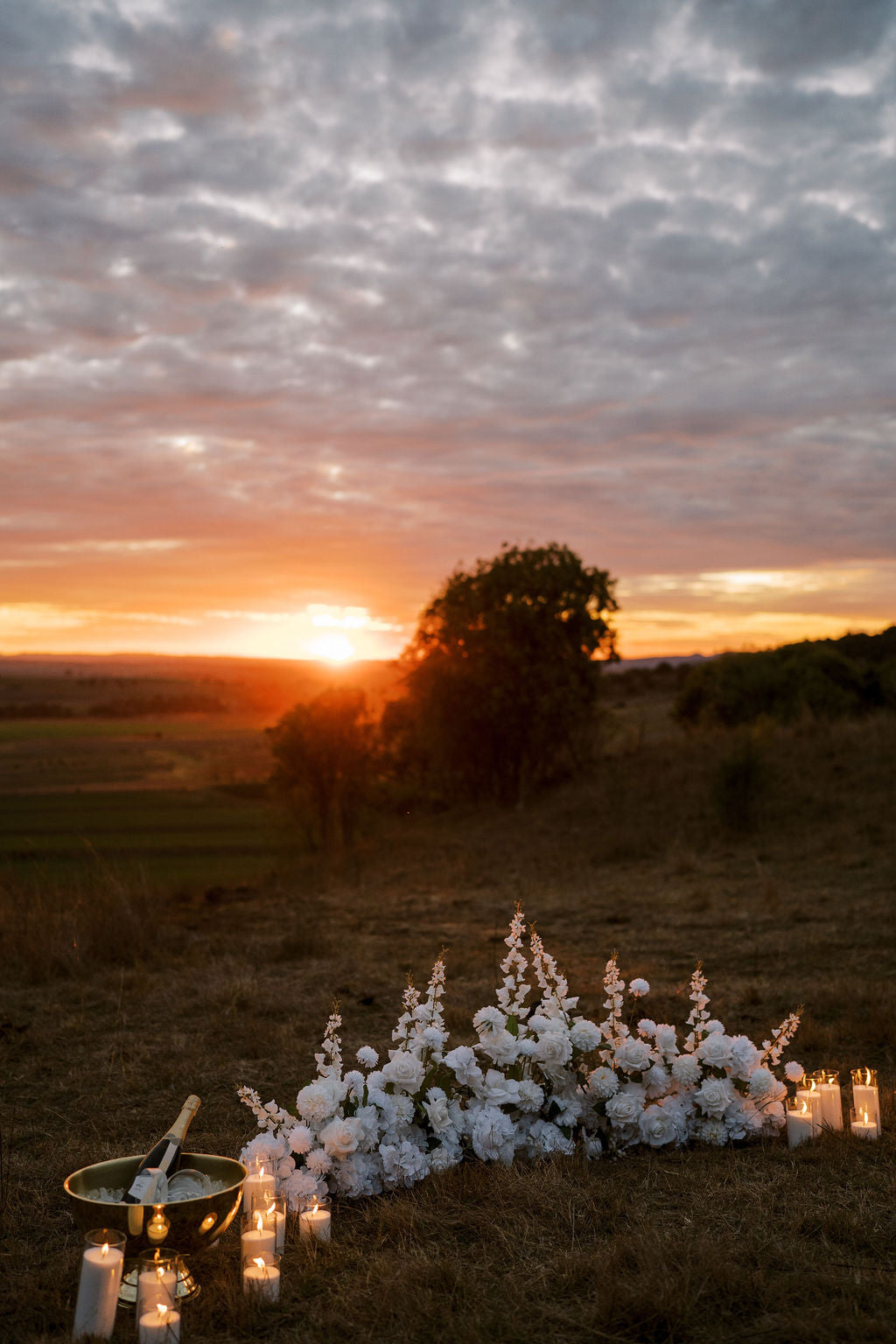 Outdoor wedding-style floral arrangement with white flowers and lit candles set in a field at sunset, with a bowl and utensils nearby and a tree silhouetted against the glowing sun.