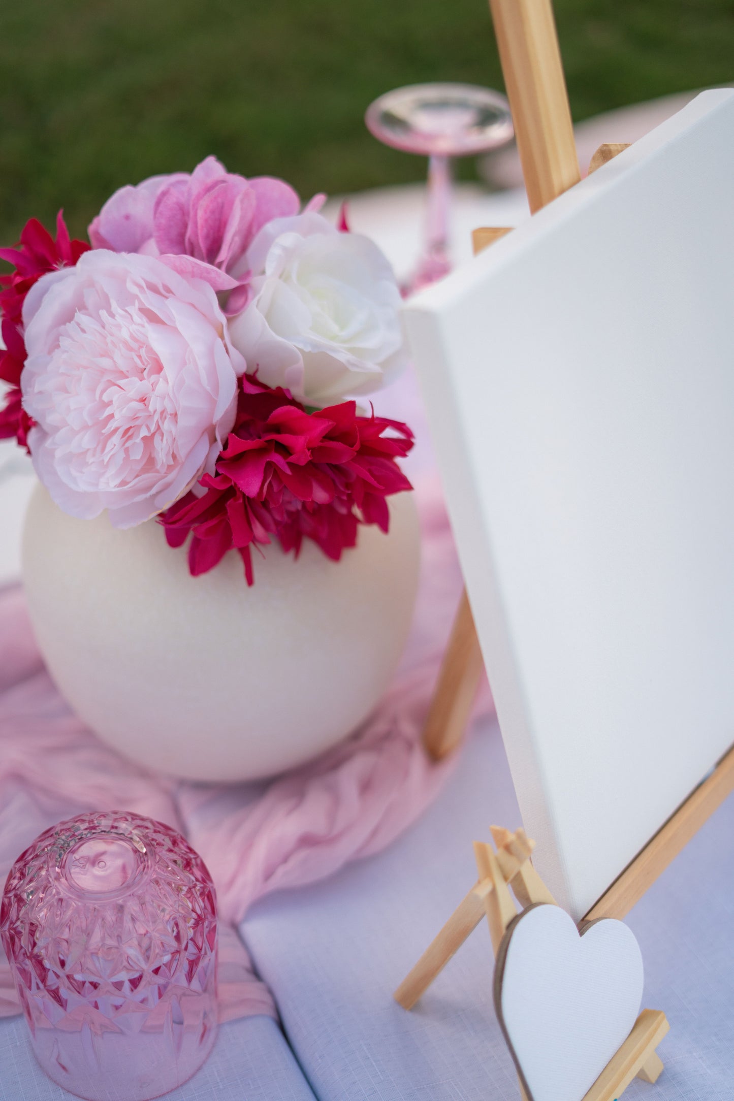 Decorative setup with pink and white flowers on a white vase, pink fabric, and heart-shaped canvas on stand.