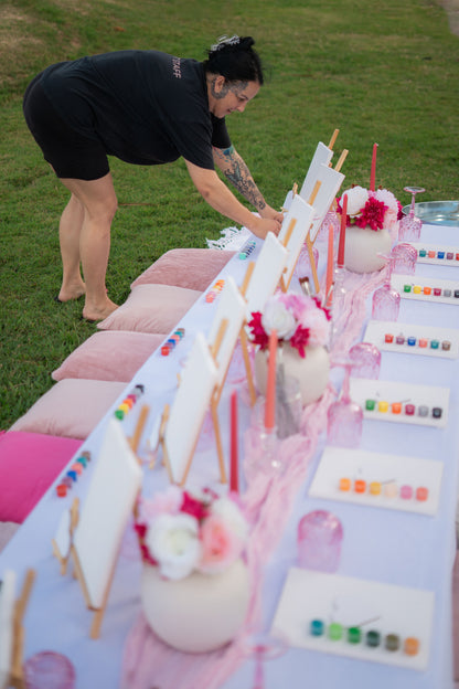 Evey setting up a sip and paint table with decorative items on a grassy background